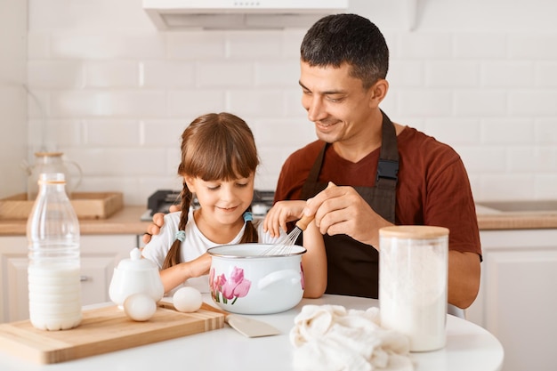 Uomo sorridente con camicia marrone e grembiule nero che aiuta la sua piccola figlia con pasta fatta in casa, regalo per la madre, famiglia in posa in cucina leggera.