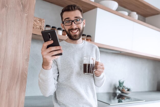 Uomo sorridente con smartphone e tazza di caffè in piedi nella sua cucina