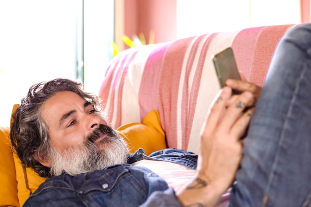 Uomo sorridente felice che utilizza un dispositivo smartphone mentre è seduto su un divano a casa Uomo maturo sdraiato sul divano leggendo i messaggi sul telefono cellulare