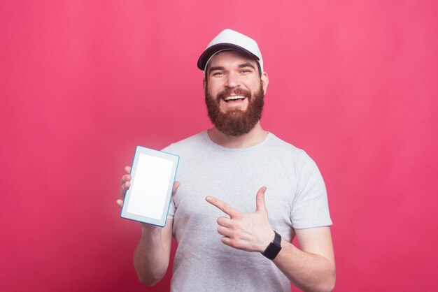 Uomo sorridente felice con la barba che indica allo schermo della compressa sopra la parete rosa