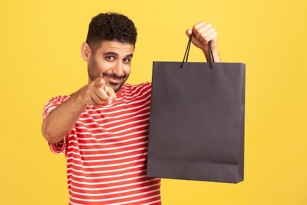 Uomo sorridente felice con la barba in maglietta a righe che punta il dito verso la macchina fotografica che tiene un sacchetto di carta nero con gli acquisti, invitandoti a fare shopping. Studio indoor girato isolato su sfondo giallo