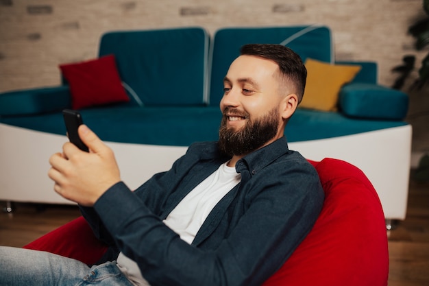 Uomo sorridente felice resto sulla poltrona in soggiorno e guardare video divertenti su smartphone.