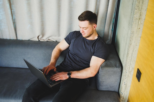 Uomo sorridente in maglietta seduto sul divano che digita sul netbook lavorando in remoto all'avvio come libero professionista guardando il laptop e sorridendo
