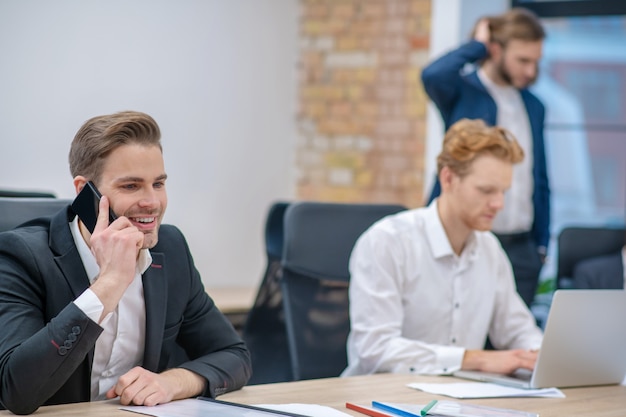 Uomo sorridente in vestito che parla sullo smartphone mentre era seduto al tavolo e lavora vicino ai colleghi in ufficio