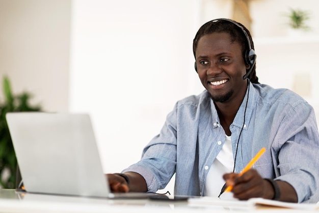 Uomo sorridente nero di formazione in linea nello studio della cuffia con il computer portatile a casa