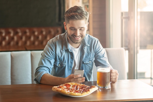 Uomo sorridente seduto al bar utilizzando smartphone e bere birra con la pizza