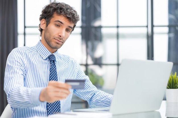 Uomo sorridente seduto in ufficio e paga con carta di credito con il suo computer portatile
