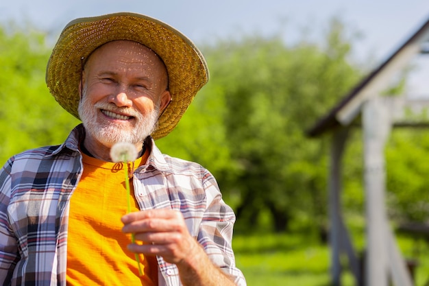 Uomo sorridente. Un uomo in pensione dai capelli grigi raggiante che indossa un cappello di paglia che sorride mentre tiene il dente di leone