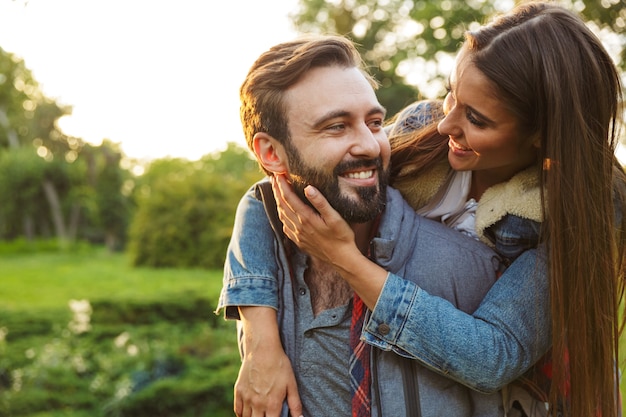 uomo sorridente vestito in abbigliamento casual che fa un giro sulle spalle alla sua ragazza nel parco verde