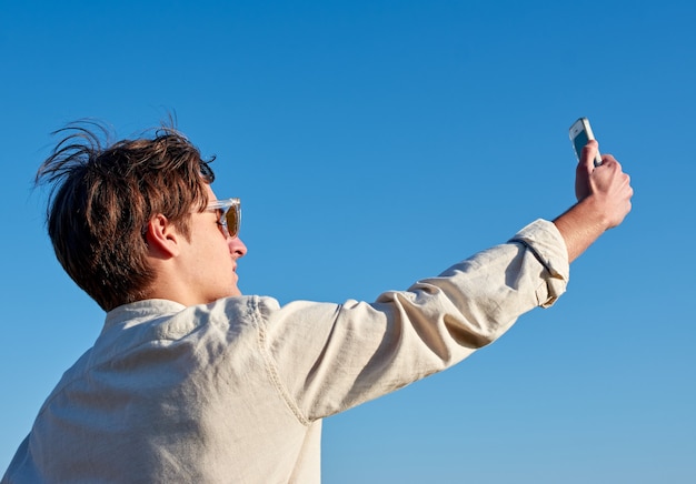 Uomo spagnolo con gli occhiali e una camicia beige che prende un selfie sul cielo blu chiaro