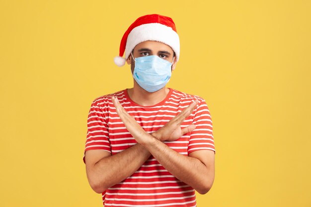 Uomo spaventato con cappello di Babbo Natale con maschera medica chirurgica sul viso in piedi incrociando le mani, che mostra il segno x che significa stop, mantenere la distanza. Studio indoor girato isolato su sfondo giallo