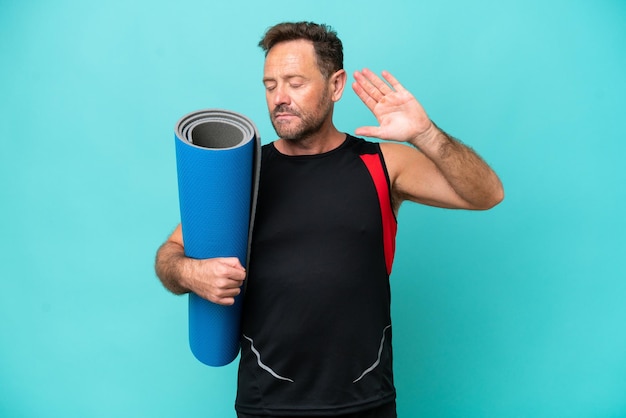 Uomo sportivo di mezza età che va alle lezioni di yoga mentre tiene un tappetino isolato su sfondo blu facendo gesto di arresto e deluso