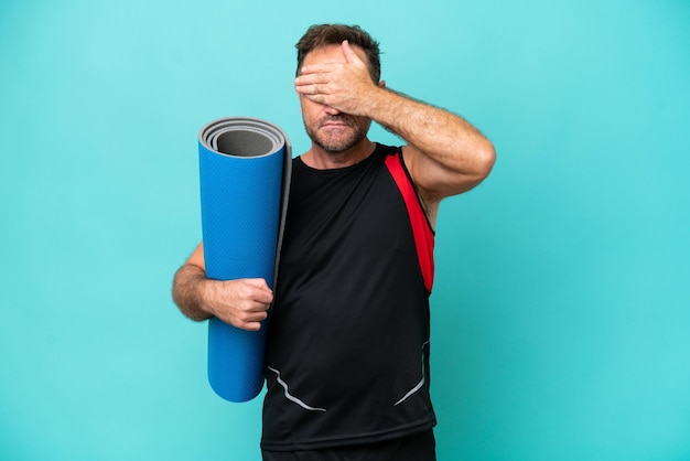 Uomo sportivo di mezza età che va alle lezioni di yoga tenendo un tappetino isolato su sfondo blu che copre gli occhi con le mani Non voglio vedere qualcosa