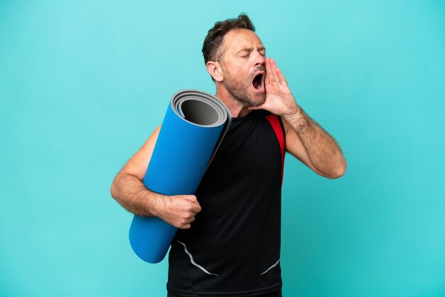 Uomo sportivo di mezza età che va alle lezioni di yoga tenendo un tappetino isolato su sfondo blu gridando con la bocca spalancata di lato