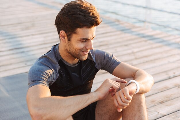 Uomo sportivo sorridente in tuta da ginnastica che si siede sulla stuoia e che esamina cronometro dopo l'allenamento sul pilastro di legno alla spiaggia nella mattina