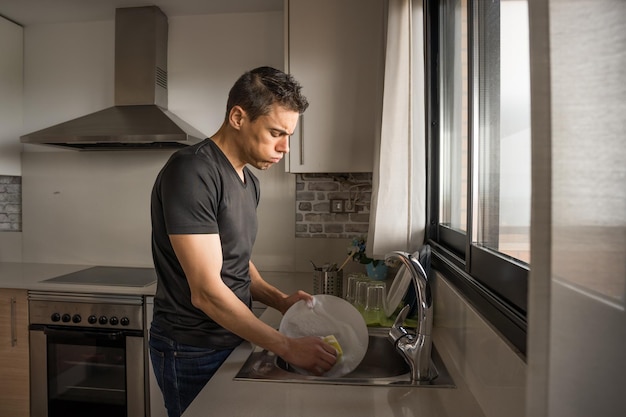 Uomo stanco che lava i piatti in cucina accanto a una finestra. Colpo medio.