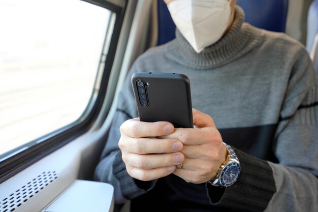 Uomo sui mezzi pubblici che utilizza l'app mobile che indossa una maschera medica Pendolare del treno che tiene cellulare con maschera protettiva obbligatoria KN95 FFP2 Focus sul telefono