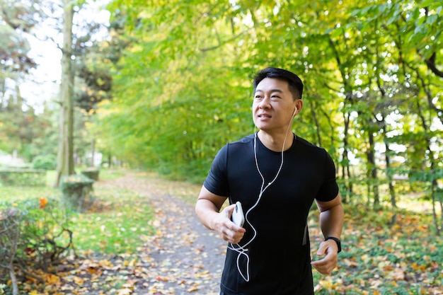 Uomo sul jogging mattutino in autunno parco sportivo asiatico in abbigliamento sportivo con cuffie e telefono nella foresta
