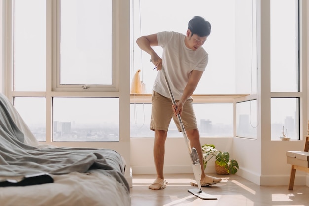 Uomo thailandese asiatico che usa la scopa per pulire il pavimento nel soggiorno dell'appartamento Uomo che fa le faccende domestiche