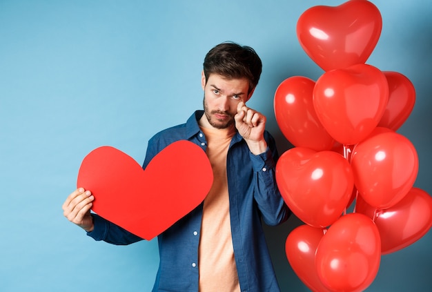 Uomo triste e affranto che piange, asciugandosi le lacrime, in piedi con cuore rosso e palloncini, rottura il giorno di San Valentino, sfondo blu.