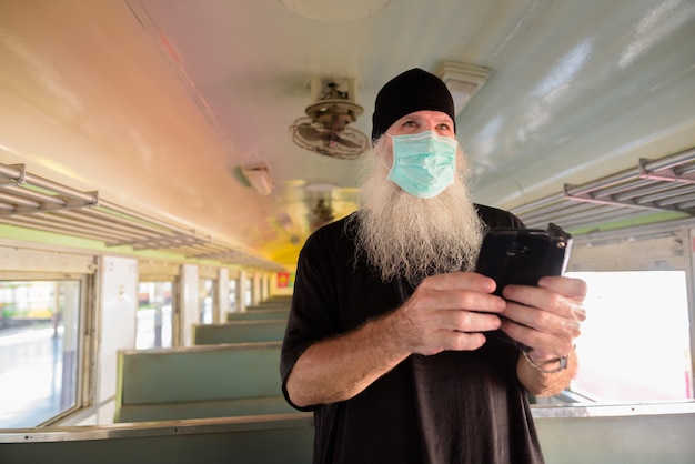 Uomo turistico barbuto maturo che pensa mentre usando telefono con la maschera per protezione dall'epidemia del virus della corona dentro il treno