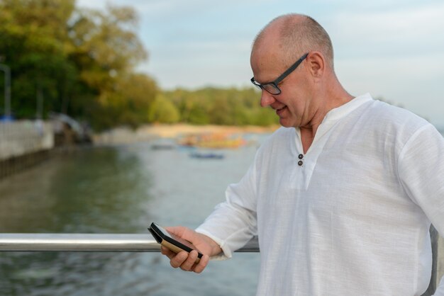 Uomo turistico bello maturo felice utilizzando il telefono contro la vista della spiaggia all'aperto