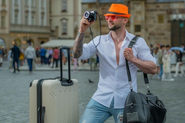 Uomo turistico in moda cappello jeans e occhiali da sole con valigia che cammina per strada Bel giovane in viaggio d'affari che cammina con i suoi bagagli per strada Uomo d'affari in viaggio