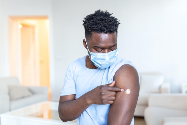 Uomo vaccinato con maschera facciale protettiva guardando il braccio con intonaco per iniezione Ritratto di uomo africano vaccinato che mostra il suo braccio
