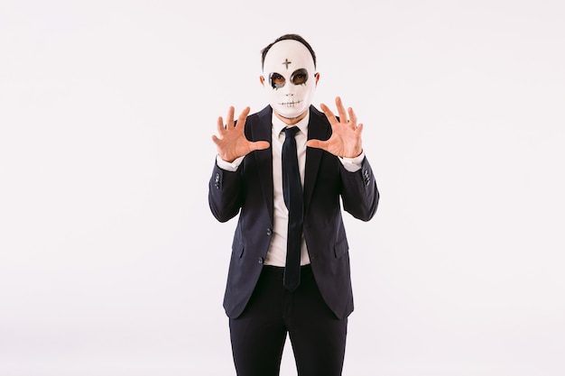Uomo vestito con giacca e cravatta, che indossa una maschera da killer con una croce sulla fronte per Halloween, che spaventa con le mani. Carnevale e festa di halloween