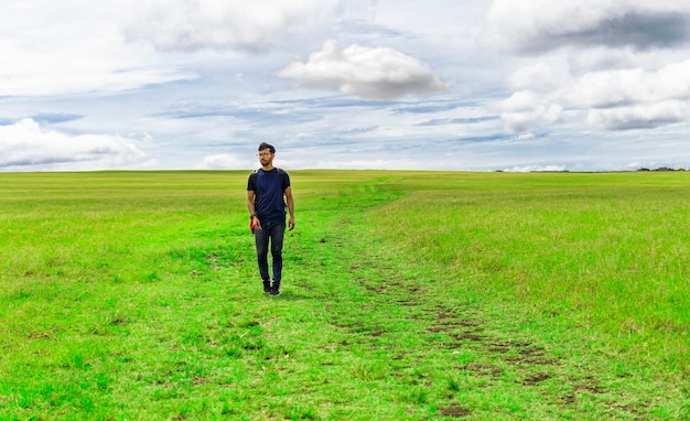 Uomo viaggiatore che cammina nella bellissima campagna Uomo viaggiatore con zaino e sacco a pelo che cammina in una campagna Paesaggio dell'uomo viaggiatore che cammina nel campo verde Stile di vita dell'uomo avventuroso nel campo