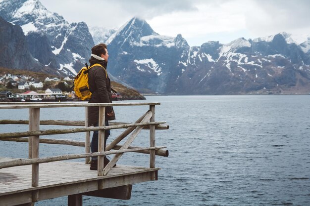 Uomo viaggiatore con uno zaino in piedi su un molo di legno sullo sfondo di montagne innevate e lago