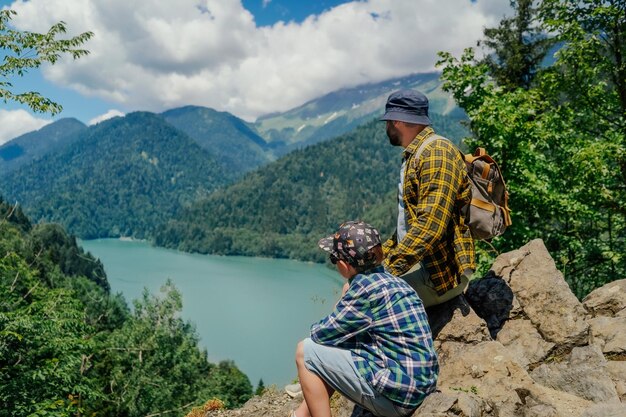 uomo viaggiatore con zaino seduto sulle rocce con figlio che gode di una splendida vista del lago blu turchese