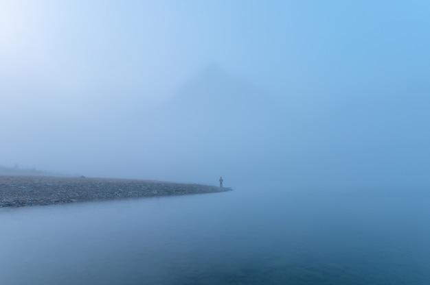 Uomo viaggiatore in piedi da solo nella nebbia blu con montagne rocciose in riva al lago al mattino