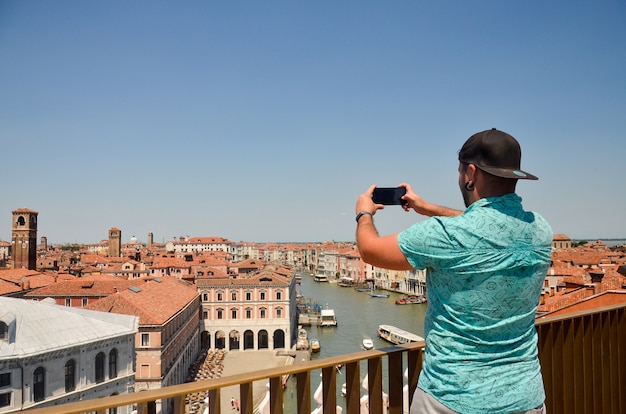 Uomo viaggio turistico in Italia. Equipaggi la fabbricazione della foto facendo uso del suo smartphone che sta e che guarda sulla cima del tetto. Vista sul canal grande. Uomo viaggio turistico in Italia.