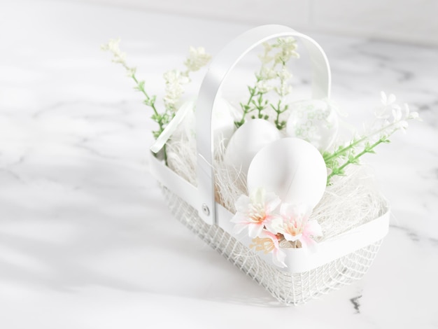 Uova bianche decorative in un cestino di metallo con fiori primaverili di uova e paglia su un tavolo di marmo