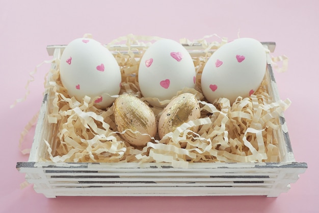 Uova bianche di Pasqua con i cuori rosa e beige in puntino nero e colpi dell'oro in un canestro su un fondo rosa.