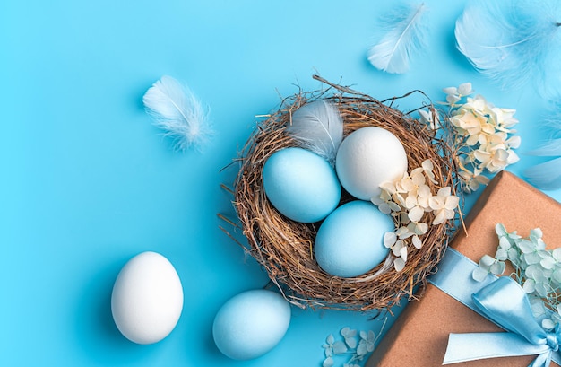 Uova bianche e blu in un nido con fiori un regalo e piume su sfondo blu Sfondo di Pasqua Spazio per la copia della vista dall'alto