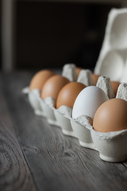 Uova di gallina marrone e un uovo bianco in un cartone su superficie di legno.