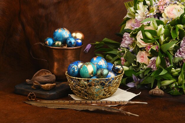 Uova di Pasqua colorate dipinte in ciotola d'oro e tazza di rame vicino a bouquet di fiori su pelle di animale rossa Stile Boho