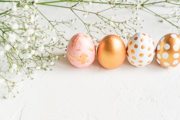 Uova di Pasqua colorate dipinte nei colori oro, bianco e rosa in fila su sfondo bianco cemento decorato con fiori di gypsophila. Tenero sfondo vacanza creativa con spazio di copia