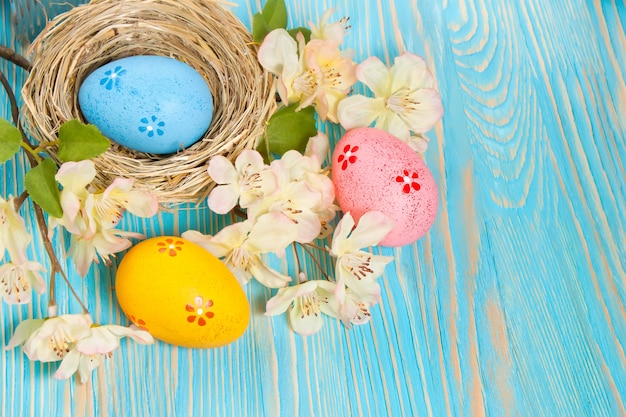 Uova di Pasqua colorate nel nido di paglia e ramo con fiori su fondo di legno blu