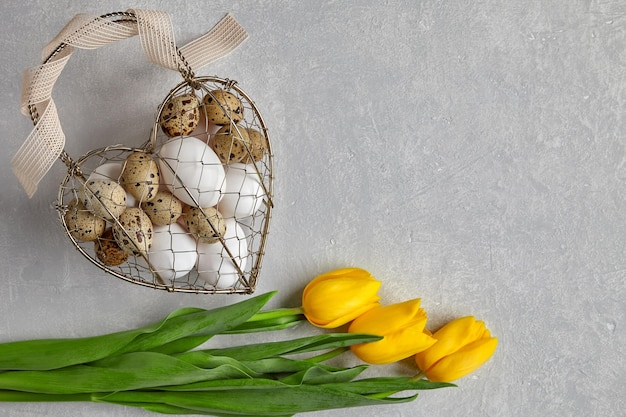 Uova di pollo e quaglia in una scatola di filo metallico intrecciato a forma di cuore legata con un nastro e tulipani gialli su uno sfondo di cemento chiaro Vista dall'alto con spazio per la copia Posa piatta
