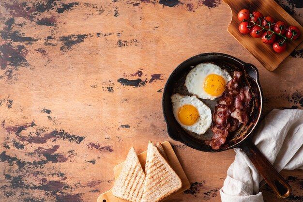 Uova fritte in padella con pomodorini secchi, pancetta e pane tostato per la colazione su un legno rustico