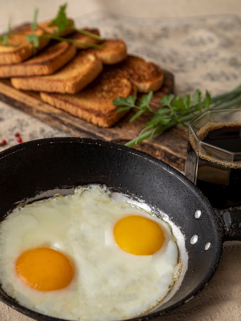 Uova fritte in una padella di ferro rustica, pane tostato su una tavola di legno e una tazza di caffè per colazione.