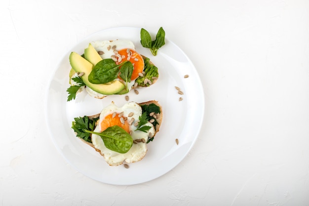 Uova fritte su un pane tostato con avocado, spinaci e semi su un piatto bianco.