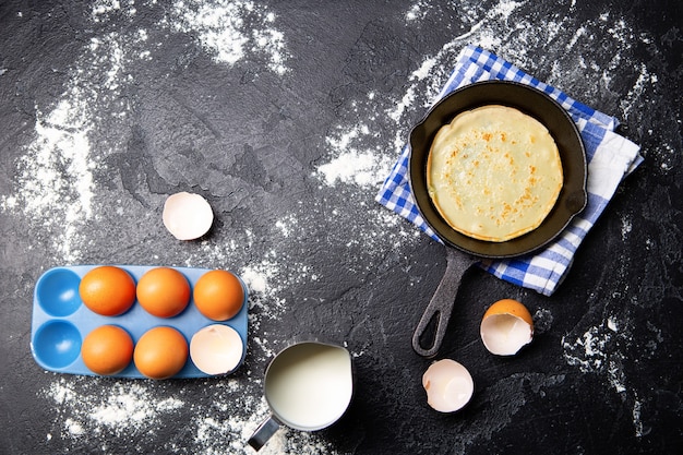 Uova, latte, padella con frittelle, asciugamani sul tavolo nero