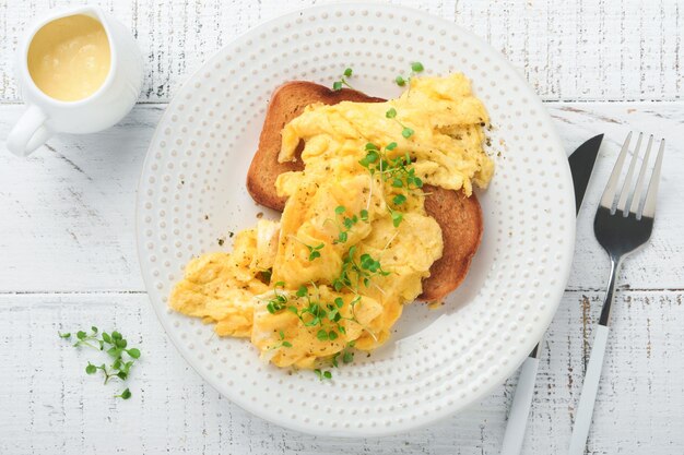 Uova strapazzate Uova strapazzate soffici e burrose su pane con ravanello microgreen e salsa olandese su piatto bianco su sfondo bianco di legno Colazione o brunch fatti in casa Vista dall'alto