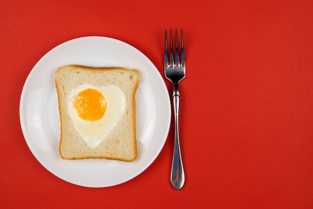 Uovo a forma di cuore in una fetta di pane di segale tostato su un piatto di ceramica bianca. Concetto di San Valentino. Adoro il design della colazione. Panino sano fatto in casa. Pranzo o colazione festivi. Copia spazio.