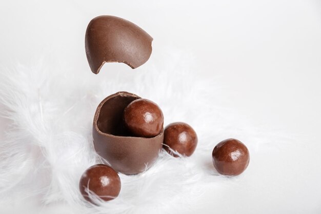 Uovo di Pasqua al cioccolato incrinato con piccole caramelle rotonde al cioccolato e guscio d'uovo volante nel nido di piume bianche