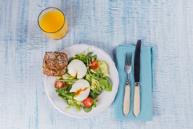 Uovo in camicia con insalata verde, pomodori, pane integrale e succo d'arancia su legno blu.